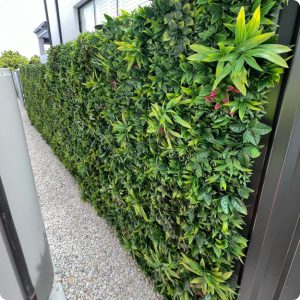 A Living Wall running along a narrow gravel pathway, featuring well-maintained foliage in various shades of green and yellow, bordered by a gray fence and a dark structure.